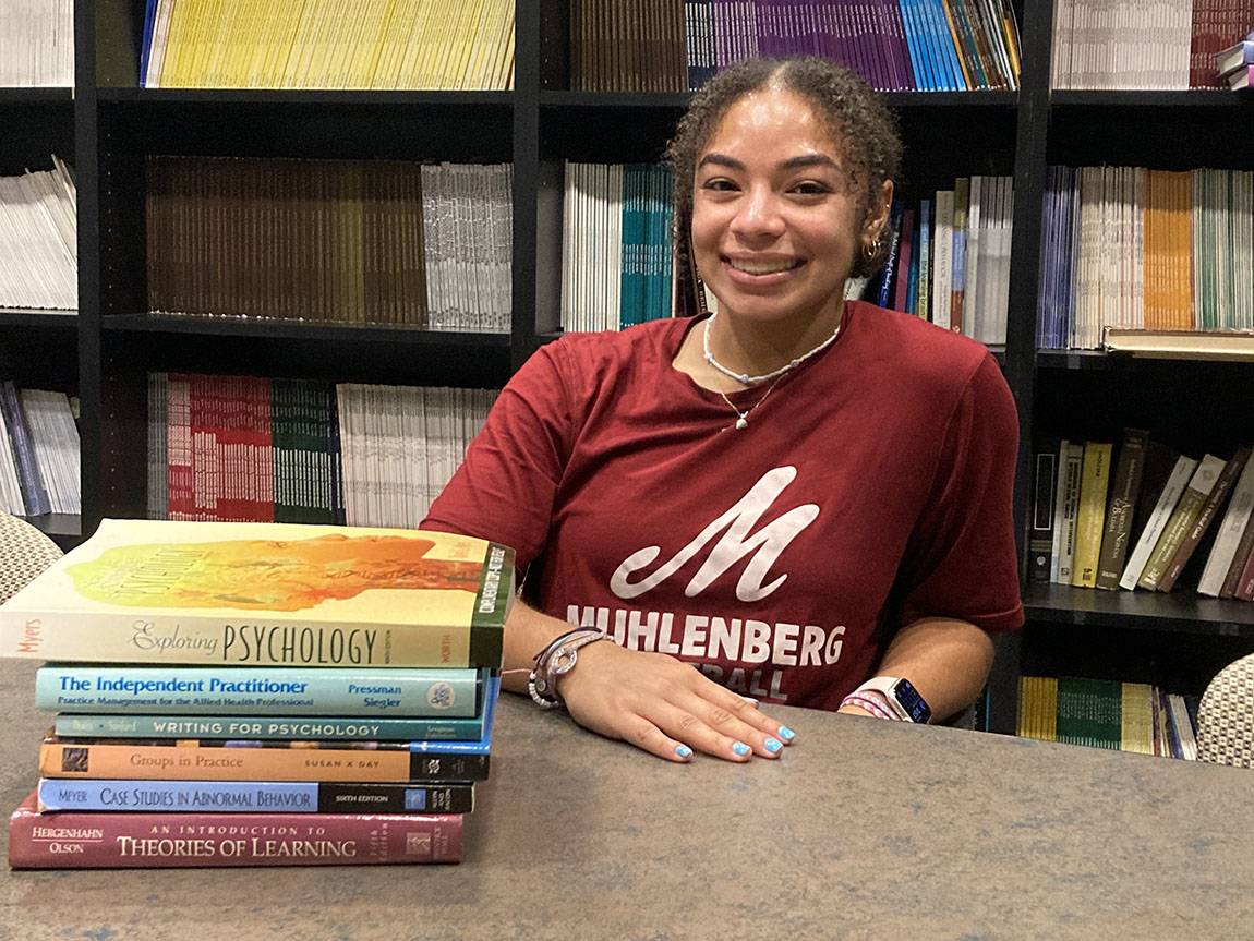 A college student smiles while sitting next to a stack of psychology books